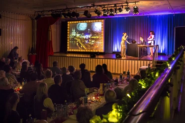 Köln ist ein Gefühl. Die Comedy Musical Dinner Show Musical Dinner Show: Auf der Bühne stehen drei Schauspieler neben einem Bild mit der Kölner Skyline. Zahlreiche Gäste sitzen an langen Banketttischen und schauen die Show