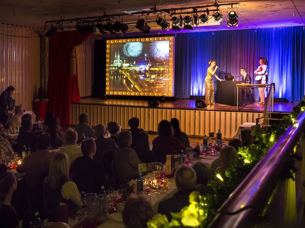 Köln ist ein Gefühl. Die Comedy Musical Dinner Show Musical Dinner Show: Auf der Bühne stehen drei Schauspieler neben einem Bild mit der Kölner Skyline. Zahlreiche Gäste sitzen an langen Banketttischen und schauen die Show
