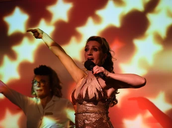 Köln ist ein Gefühl. Die Comedy Musical Dinner Show. Musicalstar Sängerin in Glitzerkleid mit Mikrofon vor leuchtendem Sternenhintergrund, Begleitperson deutet an.</p>Singer in glittering dress with microphone in front of glowing starry background, accompanying person indicates.</p> <p
