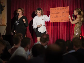 Köln ist ein Gefühl. Das kleine Gefühl. Das Dreigestirn Sängerinnen auf einer Bühne vor rotem Vorhang, eine hält ein Schild, Publikum sitzt davor.Cologne is a feeling. The small feeling. The triumvirate on stage