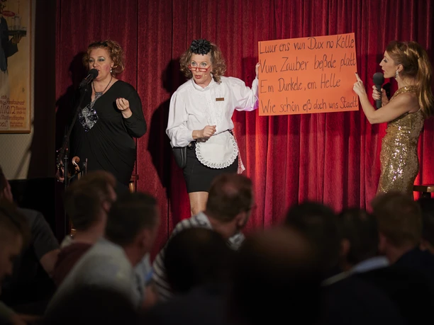 Köln ist ein Gefühl. Das kleine Gefühl. Das Dreigestirn Sängerinnen auf einer Bühne vor rotem Vorhang, eine hält ein Schild, Publikum sitzt davor.