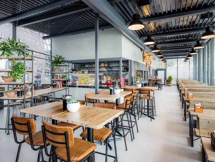Hyatt-Regency-Cologne-StickyFingers-Interior.jpg Modernes Café mit Holztischen, Stühlen, Pflanzen und großen Fenstern, das eine helle Atmosphäre bietet.</p>Modern café with wooden tables, chairs, plants and large windows, offering a bright atmosphere.
