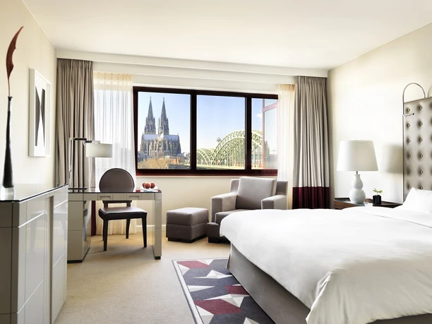Rheinblick King Zimmer Ein elegantes Hotelzimmer mit Blick auf den Kölner Dom und die Hohenzollernbrücke durch das Fenster.