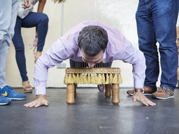 Westar. Zeigen was in uns steckt. Fakir & Feuer Ein Mann führt humorvoll Liegestütze auf einer robusten Bürste aus, umgeben von lachenden Menschen.</p>