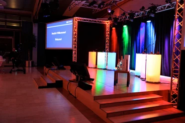 WeQuiz. Das Quizevent. Bühne Beleuchtete Bühne mit mehrfarbigen Scheinwerfern, Podium und Projektorbildschirm "Herzlich Willkommen".</p>