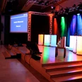 WeQuiz. Das Quizevent. Bühne Beleuchtete Bühne mit mehrfarbigen Scheinwerfern, Podium und Projektorbildschirm "Herzlich Willkommen".</p>Illuminated stage with multicolored spotlights, podium and "Welcome" projector screen.</p> <p