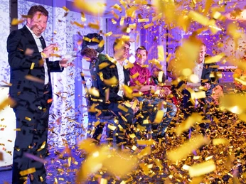 WeQuiz. Das Quizevent. Siegerehrung Ein Mann in Anzug lächelt, während goldener Konfetti-Regen bei einem Event fällt.A man in a suit smiles as golden confetti falls at an event.