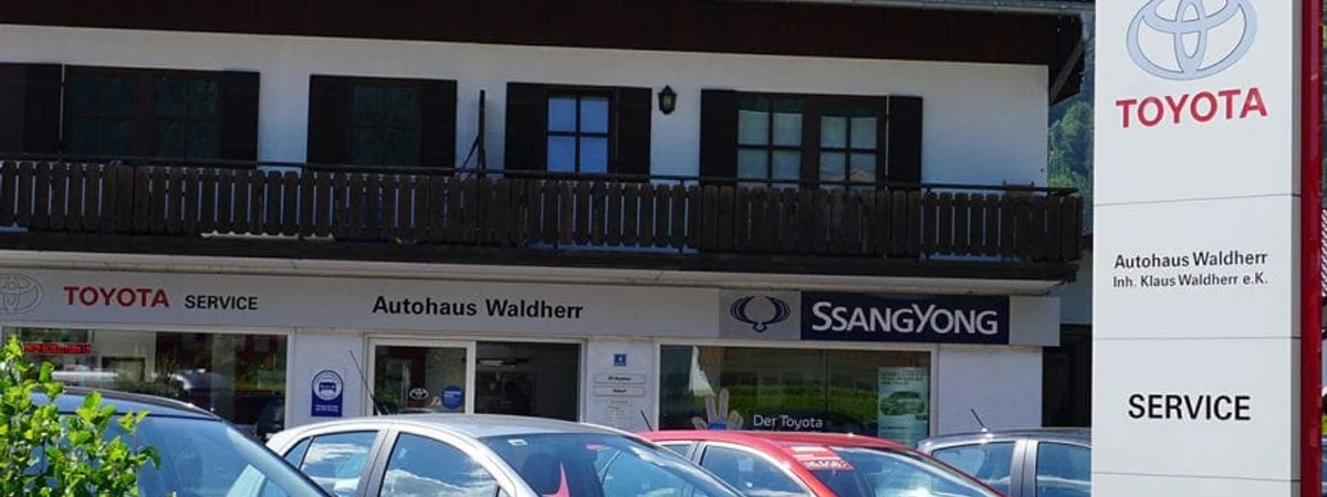Autohaus Waldherr Farchant Haus mit Aufschrift Toyota, Autohaus Waldherr SsangYong. Vor dem Haus stehen 3 Autos und eine Anzeigetafel mit der Aufschrift Toyota, Autothaus Waldherr und Service