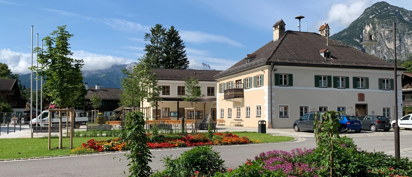 Rathaus Farchant, davor Blumenbeet im Hintergrund Bäume, der Kramer und weiter hinten das Wettersteingebirge mit Zugspitze