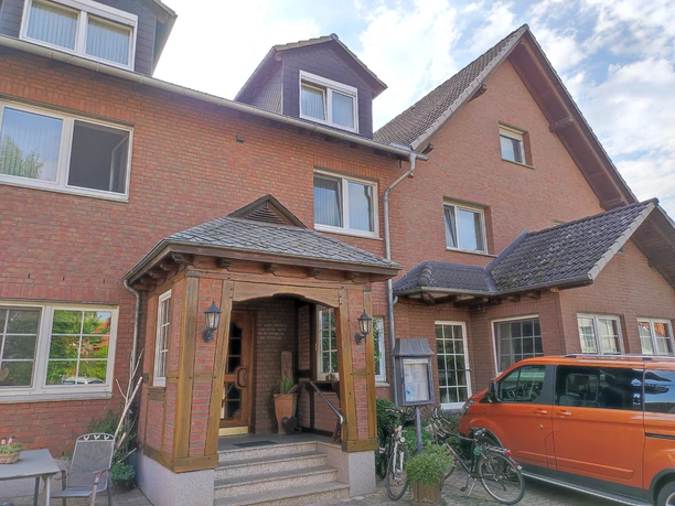 Rot verklinkertes Gasthaus mit Dreiecksgauben, Eingangsstufen und parkendem orangenen Auto davor.