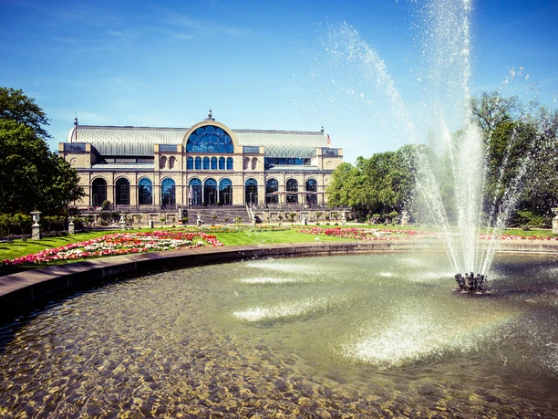 Flora Köln, Außenansicht mit Brunnen Historische Halle mit großem Glasfenster, umgeben von Parkanlagen und einem brunnengekrönten Teich.</p>