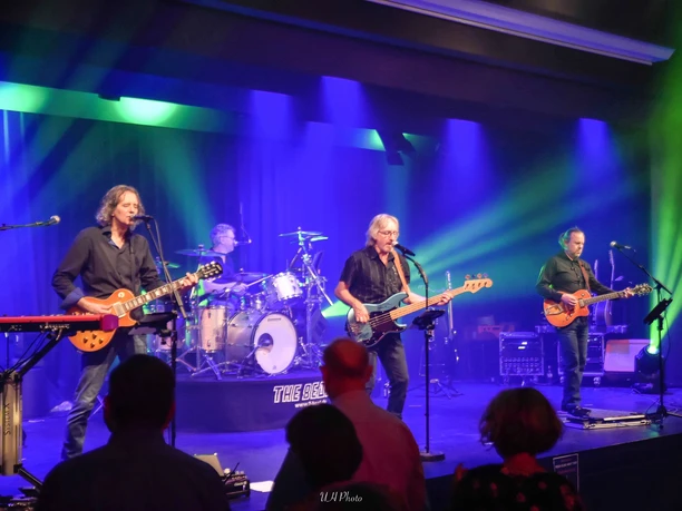 -c-the beat.jpg Eine vierköpfige Band spielt auf einer lebhaft beleuchteten Bühne vor einem begeisterten Publikum.