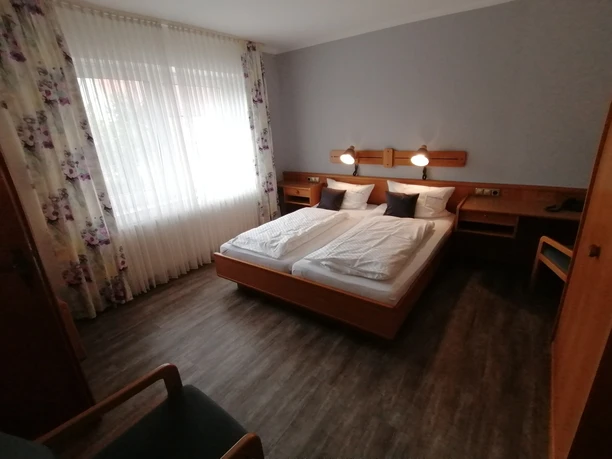 Doppelzimmer Schierbrucher Krug.jpg Helles, modern eingerichtetes Doppelzimmer mit zwei Betten, floral gemusterten Vorhängen und Schreibtisch.