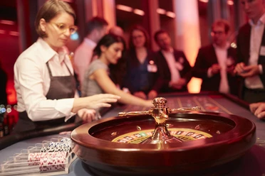 Casino Vinophil. Rebsorten Roulette. Dieter Jacobi.jpg Roulettetisch in edlem Casino, im Hintergrund Gäste und Croupier unter warmem Licht.