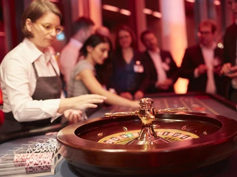 Casino Vinophil. Rebsorten Roulette. Dieter Jacobi.jpg Roulettetisch in edlem Casino, im Hintergrund Gäste und Croupier unter warmem Licht.Roulette table in a classy casino, in the background guests and croupier under warm light.