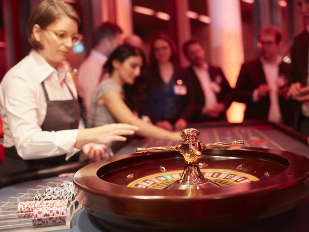 Casino Vinophil. Rebsorten Roulette. Dieter Jacobi.jpg Roulettetisch in edlem Casino, im Hintergrund Gäste und Croupier unter warmem Licht.