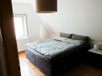 2023-06-HausGörlich-21.jpg Helles Schlafzimmer mit Doppelbett, Bettwäsche in Grautönen, Holzboden, Stehlampe, kleinem Nachttisch.