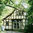 2023-06-HausGörlich-49 Kopie.jpg Ein Fachwerkhaus mit großen Fenstern liegt umgeben von üppigem Grün in einem friedlichen Wald.