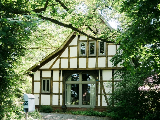 2023-06-HausGörlich-49 Kopie.jpg Ein Fachwerkhaus mit großen Fenstern liegt umgeben von üppigem Grün in einem friedlichen Wald.