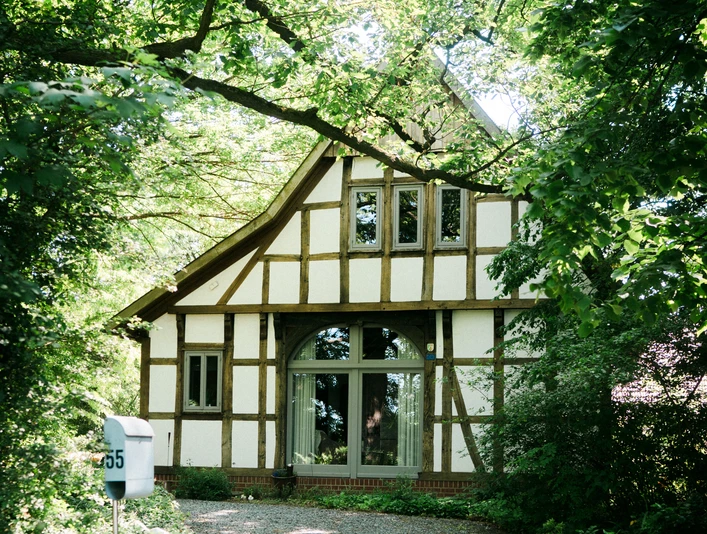 2023-06-HausGörlich-49 Kopie.jpg Ein Fachwerkhaus mit großen Fenstern liegt umgeben von üppigem Grün in einem friedlichen Wald.