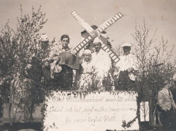 tengern-finkmanns-muehle-02.jpg Schwarz-Weiß-Foto einer Gruppe in traditioneller Kleidung, umgeben von Laubzweigen und Windmühle.