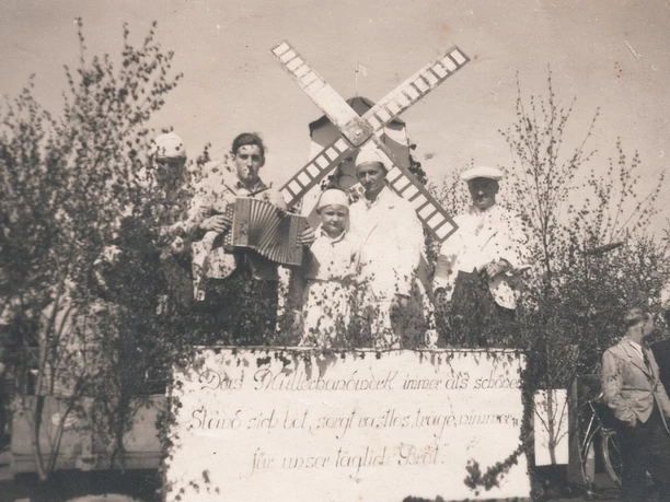 tengern-finkmanns-muehle-02.jpg Schwarz-Weiß-Foto einer Gruppe in traditioneller Kleidung, umgeben von Laubzweigen und Windmühle.