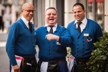 Typical Köbesse Three Köbesse, waiters in traditional blue clothing with ties, laughing heartily. They radiate the kind of friendly and welcoming atmosphere often found in breweries. Their cheerfulness conveys a feeling of conviviality and hospitality.