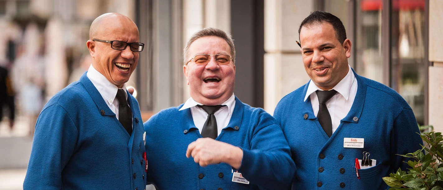 Typical Köbesse Three Köbesse, waiters in traditional blue clothing with ties, laughing heartily. They radiate the kind of friendly and welcoming atmosphere often found in breweries. Their cheerfulness conveys a feeling of conviviality and hospitality.