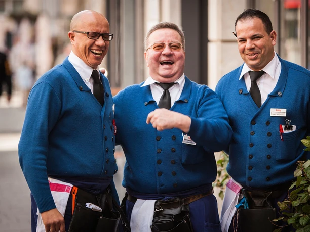 Typical Köbesse Three Köbesse, waiters in traditional blue clothing with ties, laughing heartily. They radiate the kind of friendly and welcoming atmosphere often found in breweries. Their cheerfulness conveys a feeling of conviviality and hospitality.