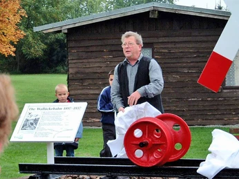 Wallücker Wilhelm Mann steht neben rot-weißem Modell eines Mühlrads, umgeben von Menschen, grüner Wiese, Holzhütte im Hintergrund.