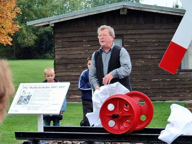 Wallücker Wilhelm Mann steht neben rot-weißem Modell eines Mühlrads, umgeben von Menschen, grüner Wiese, Holzhütte im Hintergrund.