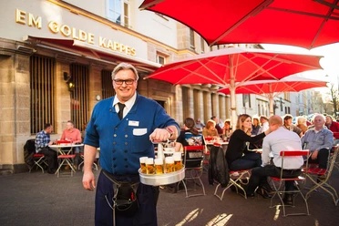 Früh Em Golde Kappes Auf dem Bild ist ein Kellner zu sehen, der eine blaue Jacke trägt und einen Kranz mit Biergläsern in der Hand hält. Er lächelt freundlich in die Kamera. Im Hintergrund sitzen Gäste an Tischen mit roten Sonnenschirmen vor einem Gebäude, auf dem "Em Golde Kappes" steht. Die Szene vermittelt eine gesellige Stimmung in einer Außengastronomie.