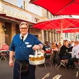 Früh Em Golde Kappes Auf dem Bild ist ein Kellner zu sehen, der eine blaue Jacke trägt und einen Kranz mit Biergläsern in der Hand hält. Er lächelt freundlich in die Kamera. Im Hintergrund sitzen Gäste an Tischen mit roten Sonnenschirmen vor einem Gebäude, auf dem "Em Golde Kappes" steht. Die Szene vermittelt eine gesellige Stimmung in einer Außengastronomie.The picture shows a waiter wearing a blue jacket and holding a wreath of beer glasses. He smiles friendly into the camera. In the background, guests are sitting at tables with red parasols in front of a building that reads "Em Golde Kappes". The scene conveys a convivial atmosphere in an outdoor restaurant.