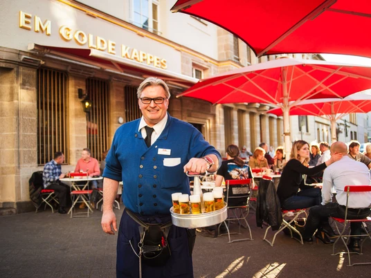 Früh Em Golde Kappes Auf dem Bild ist ein Kellner zu sehen, der eine blaue Jacke trägt und einen Kranz mit Biergläsern in der Hand hält. Er lächelt freundlich in die Kamera. Im Hintergrund sitzen Gäste an Tischen mit roten Sonnenschirmen vor einem Gebäude, auf dem "Em Golde Kappes" steht. Die Szene vermittelt eine gesellige Stimmung in einer Außengastronomie.The picture shows a waiter wearing a blue jacket and holding a wreath of beer glasses. He smiles friendly into the camera. In the background, guests are sitting at tables with red parasols in front of a building that reads "Em Golde Kappes". The scene conveys a convivial atmosphere in an outdoor restaurant.
