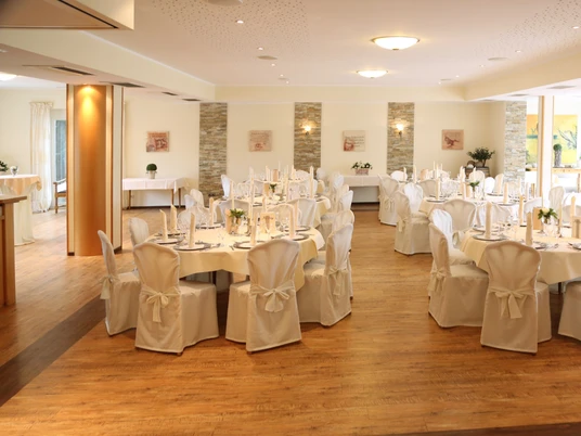 Eleganter Festsaal mit weiß gedeckten Tischen, Kerzenleuchtern und großen Fenstern mit Gartenblick.