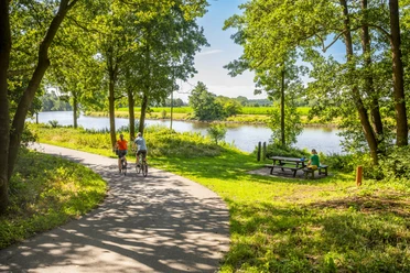 Rastplatz in Emen Zwei Radfahrende auf schattigem Weg am Kanal, nebenan Picknickbank unter Bäumen in Sommerlandschaft.