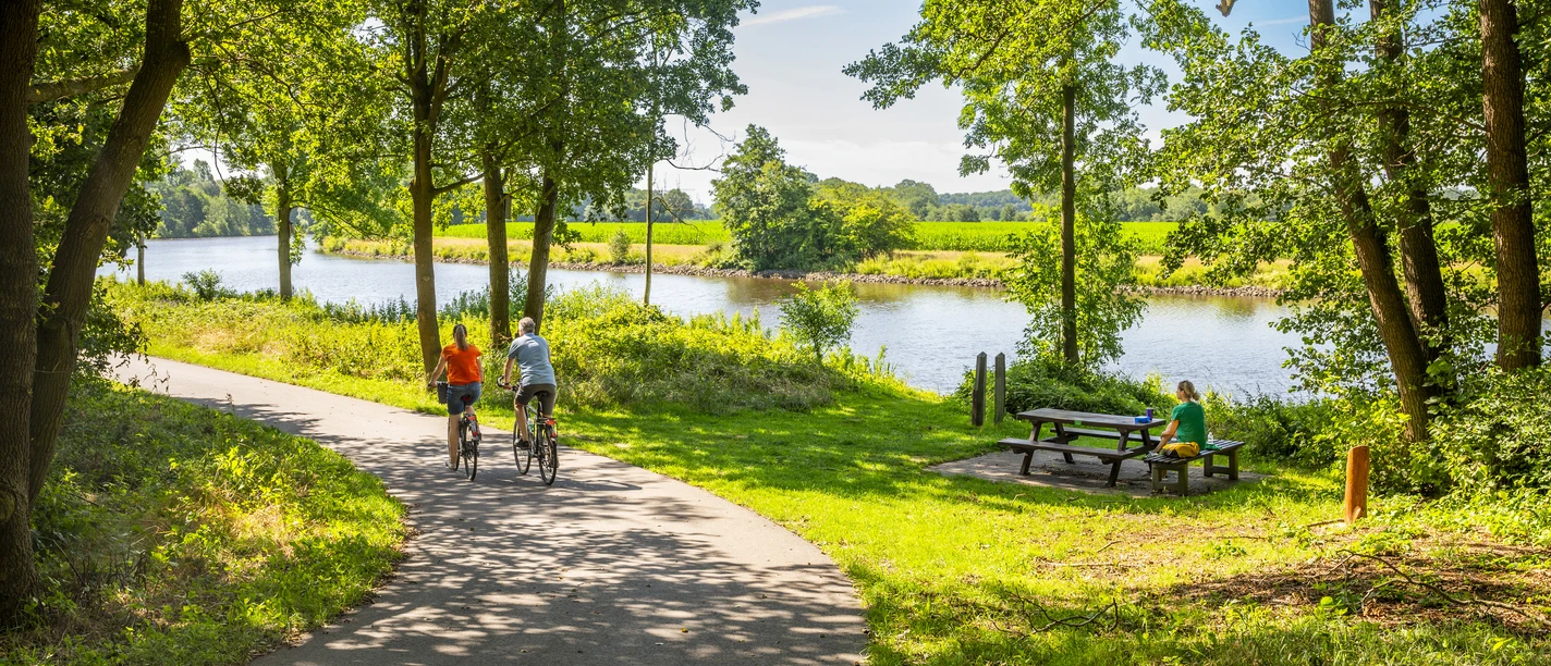 Rastplatz in Emen Zwei Radfahrende auf schattigem Weg am Kanal, nebenan Picknickbank unter Bäumen in Sommerlandschaft.