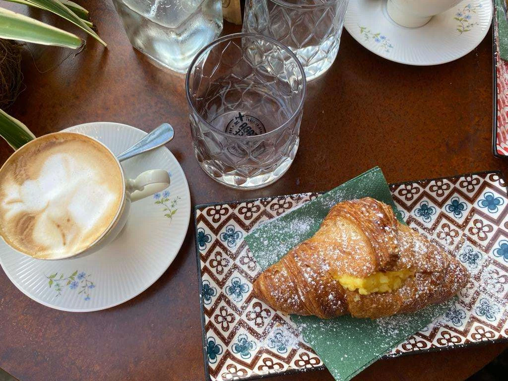 kaffeetisch.jpg Capuccino mit Schaum und Croissant auf Teller mit Puderzucker, auf Tisch mit Wasser und Gläsern.