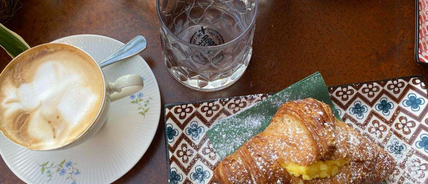 kaffeetisch.jpg Capuccino mit Schaum und Croissant auf Teller mit Puderzucker, auf Tisch mit Wasser und Gläsern.