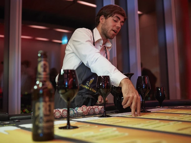 Hopfen & Malz. Bier-Poker Tisch. Dieter Jacobi.jpg Mann im weißen Hemd erklärt Karten an einem Tisch mit Weingläsern und Bierflasche.</p>