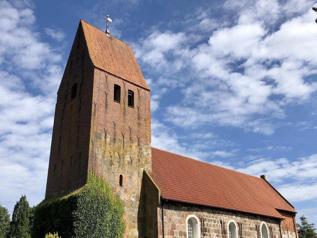 IMG_2933.jpg Backsteinkirche mit hohem Turm, umgeben von blauem Himmel und grünen Sträuchern.