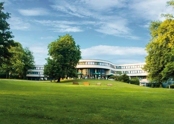 Best Western Plus Parkhotel Velbert Modernes Hotelgebäude umgeben von einer weitläufigen, grünen Parklandschaft mit Sitzbänken.