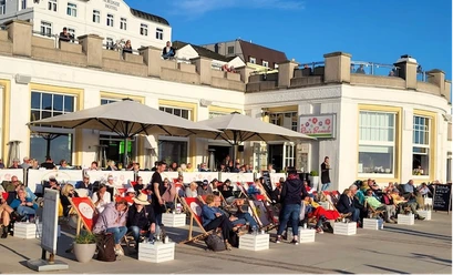 RiasBeachBorkum.png Menschen entspannen auf Liegestühlen vor einem Café bei sonnigem Wetter in Borkum.