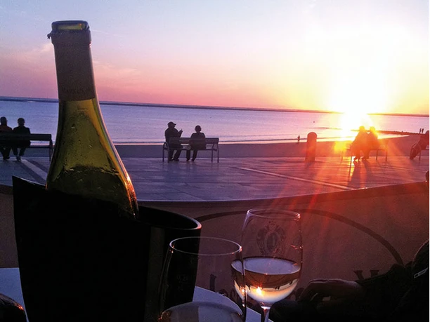 A romantic sunset at Rias Beach, with two glasses of white wine and a bottle in front of the sea on a table, people sitting on the shore enjoying the view.