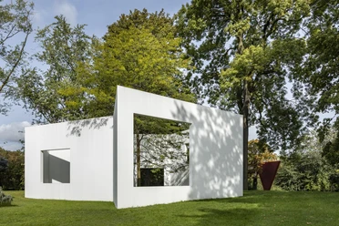 Sculpture park White, rectangular sculpture in a green park, surrounded by tall trees, showing a window.
