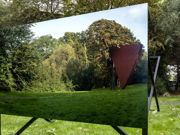 Sculpture park The picture shows an outdoor sculpture in the Cologne Sculpture Park, embedded in lush greenery and autumnal foliage. A rectangular, reflective surface can be seen in the foreground, mirroring the surrounding nature and an abstract, triangular red sculpture.