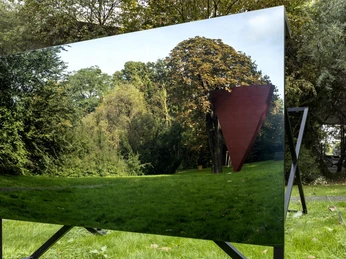 Skulpturenpark Das Bild zeigt eine Außenskulptur im Skulpturenpark Köln, eingebettet in üppiges Grün und herbstliches Laub. Im Vordergrund ist eine rechteckige, reflektierende Fläche zu sehen, die die umgebende Natur und eine abstrakte, dreieckige rote Skulptur spiegelt.The picture shows an outdoor sculpture in the Cologne Sculpture Park, embedded in lush greenery and autumnal foliage. A rectangular, reflective surface can be seen in the foreground, mirroring the surrounding nature and an abstract, triangular red sculpture.