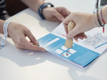 SINNfonie. Ein Parcours durch die Welt der Sinne. Laufkarte. Person erhält Dokumentenstempel auf Broschüre, während eine andere zeigt, wo gestempelt wird.</p>Person receives document stamps on brochure, while another shows where to stamp.</p> <p