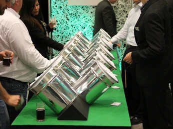 SINNfonie. Eine Parcours durch die Welt der Sinne. Taststation Stahlkessel in zwei Reihen mit Papier an einem Messestand, umgeben von Menschen in Anzügen.SINNfonie. A tour through the world of the senses. Touch station