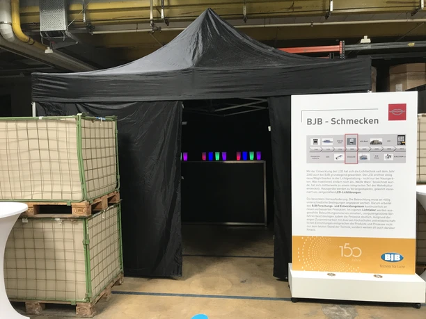 SINNfonie. Ein Parcours durch die Welt der Sinne. Schmeckstation Zelt auf Messe mit BJB-Stand, beleuchteten Displays und Informationsplakat zu LED-Technologie.